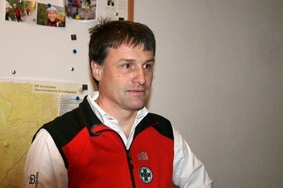 Ein Mann in einem roten und weißen Jacke mit einem Kreuz-Symbol steht vor einer Pinnwand mit Fotos und einer Karte.