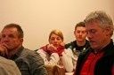 Eine Gruppe von Menschen sitzt in einem Raum, wobei ein Mann eine Frau ansieht, die nach unten schaut. Ein anderer Mann schaut etwas vor sich an.