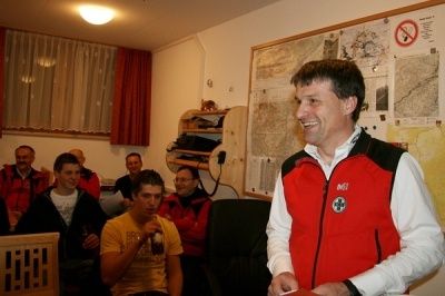 Ein Mann in einer roten Weste lächelt, während er in einem Raum steht, in dem andere Leute auf Stühlen sitzen.