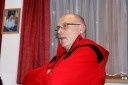 Ein älterer Mann mit Brille und rotem Mantel sitzt in einem Raum mit roten Vorhängen und einem Bilderrahmen an der Wand.