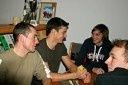 Vier Personen sitzen um einen Tisch herum und unterhalten sich. Eine Person hält eine Karte, während andere zusehen. Im Hintergrund sind ein Bücherregal und ein Bilderrahmen zu sehen.
