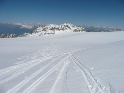 Eine verschneite Berglandschaft mit Spuren, die zu einem Gipfel führen, unter einem klaren blauen Himmel.
