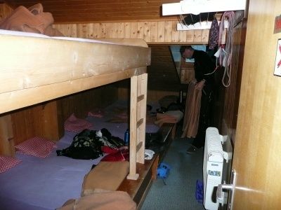 Ein kleiner Raum mit Holzwänden und Etagenbetten, eine Person steht in der Nähe der Tür und schaut etwas an.
