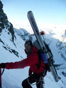 Ein Bergsteiger in einem roten Jackett steigt einen schneebedeckten Berg hinauf und trägt Skier und einen Rucksack auf dem Rücken.