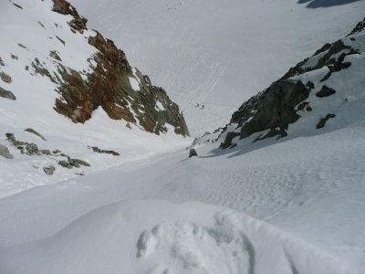 Ein schneebedeckter Berg mit einem Skifahrer, der einen steilen Hang hinunterfährt, umgeben von felsigen Klippen und einem klaren blauen Himmel.