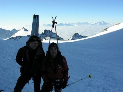 Zwei Männer stehen auf einem schneebedeckten Berggipfel, beide mit Skiern und Winterausrüstung ausgestattet, lächeln für die Kamera.