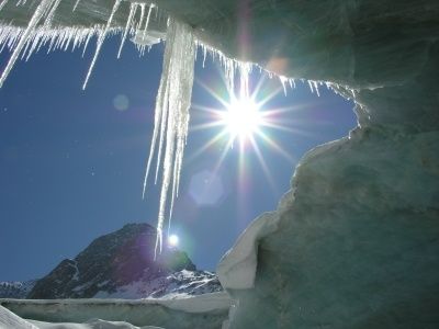 Ein Blick von innen aus einer Eishöhle mit Eiszapfen, die von der Decke hängen, und der Sonne, die hell durch ein Loch im Eis scheint.