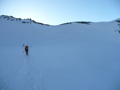 Ein einzelner Skifahrer bewegt sich auf einem verschneiten Berg und trägt Skistöcke und einen Rucksack, während ein klarer blauer Himmel über ihm ist.