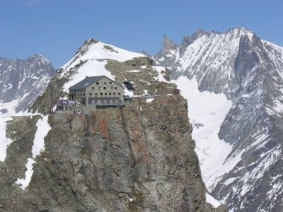 Eine Berghütte auf einem Felsvorsprung mit schneebedeckten Bergen im Hintergrund bei blauem Himmel.