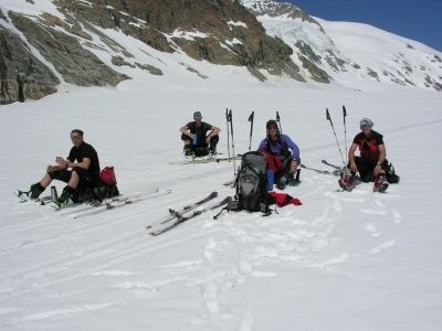 Vier Skifahrer ruhen sich auf einem verschneiten Berg aus, jeder mit Skiern und Stöcken, umgeben von Rucksäcken und Skiausrüstung.