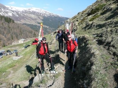 Eine Gruppe von Menschen in roten Jacken wandert mit Skistöcken einen Bergpfad hinauf. Der Pfad ist von Gras und Felsen umgeben. In der Ferne gibt es Berge und einen klaren Himmel.