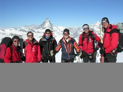 Eine Gruppe von Bergsteigern steht auf einem verschneiten Berg, alle in roten und blauen Jacken, mit Skiern und Rucksäcken. Der Matterhorn-Gipfel ist im Hintergrund zu sehen.