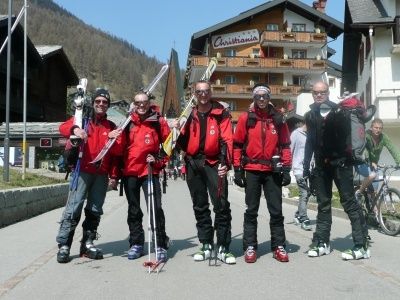 Fünf Männer in roten Jacken und Skiausrüstung stehen auf einer Straße mit Bergen im Hintergrund. Sie halten Skistöcke und Skier. Ein Gebäude mit Balkonen steht hinter ihnen.
