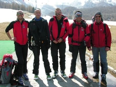 Fünf Männer in roten Jacken und Winterausrüstung stehen gemeinsam auf einem verschneiten Pfad, mit Bergen im Hintergrund. Sie tragen alle Sonnenbrillen und scheinen für ein Foto zu posieren.