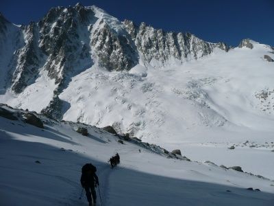Drei Bergsteiger steigen einen verschneiten Berg hinauf, ausgestattet mit Rucksäcken und Skistöcken. Der Berg ist schneebedeckt, mit sichtbaren Felsen und Eis. Der Himmel ist klar und blau.