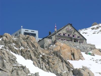 Eine Berghütte mit einer Flagge auf dem Dach, umgeben von Felsen und Schnee. Die Hütte hat viele Fenster und einen Schornstein. Ein kleineres Gebäude befindet sich in der Nähe.