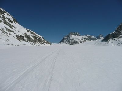 Eine weite, verschneite Landschaft mit klarem blauem Himmel und rauen Bergen im Hintergrund. Reifenspuren durchziehen den Schnee und führen zu den Bergen.