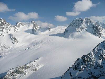 Luftaufnahme einer verschneiten Bergkette mit scharfen Gipfeln unter einem klaren blauen Himmel mit verstreuten Wolken.