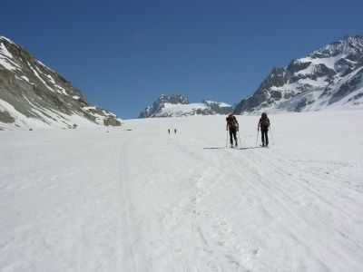 Zwei Personen üben Langlauf auf einem verschneiten Berg, mit entfernten Figuren und majestätischen Gipfeln im Hintergrund.