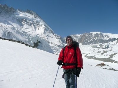 Ein Mann in einem roten Jackett und Hut steht auf einem verschneiten Berg, hält Skistöcke in der Hand, mit einem Gletscher und schneebedeckten Bergen im Hintergrund.