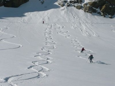 Drei Skifahrer erkunden einen verschneiten Berg und hinterlassen deutliche Spuren im Schnee. Die Landschaft ist rau mit sichtbaren Felsen.