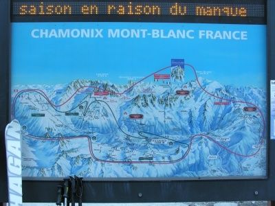 Ein digitales Kartendisplay zeigt die Skipisten von Chamonix Mont-Blanc in Frankreich, mit verschiedenen in Rot und Blau markierten Wegen und Pfaden. Das Display ist auf einem schwarzen Rahmen montiert, mit einem weißen Snowboard links sichtbar.