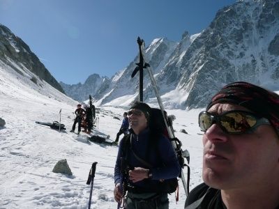 Zwei Männer mit Sonnenbrillen und Rucksäcken wandern einen verschneiten Berg hinauf, mit einer Gruppe im Hintergrund.