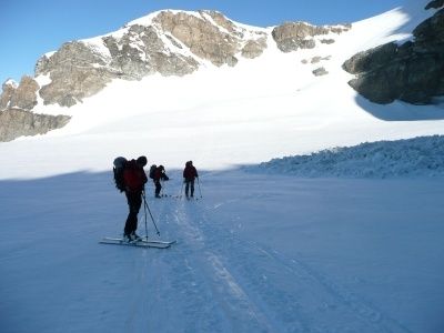 Drei Skifahrer durchqueren einen verschneiten Berg, jeder mit Rucksäcken und Skistöcken ausgestattet. Der Schnee ist dick und unberührt, mit einem Hintergrund aus felsigen Bergen.