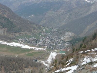 Luftaufnahme eines Bergdorfes, umgeben von schneebedeckten Hängen und grünen Feldern, mit entfernten Bergen im Hintergrund.