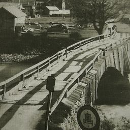 Ein Schwarz-Weiß-Foto einer Holzbrücke über einen Fluss mit einem Schild an der Seite. Im Hintergrund sind Gebäude und Autos zu sehen.