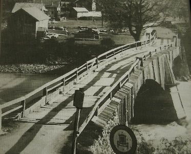 Ein Schwarz-Weiß-Foto einer Holzbrücke über einen Fluss mit einem Schild an der Seite. Im Hintergrund sind Gebäude und Autos zu sehen.