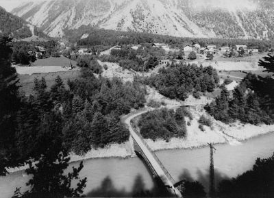 Ein Schwarz-Weiß-Foto einer bergigen Landschaft mit einer Brücke über einem Fluss und einem kleinen Dorf, das zwischen den Bäumen liegt.