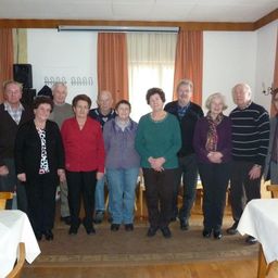 Eine Gruppe älterer Menschen steht zusammen in einem Raum und lächelt für ein Foto. Sie sind warm gekleidet und einige tragen Brillen. Der Raum hat Holzböden und weiße Wände.