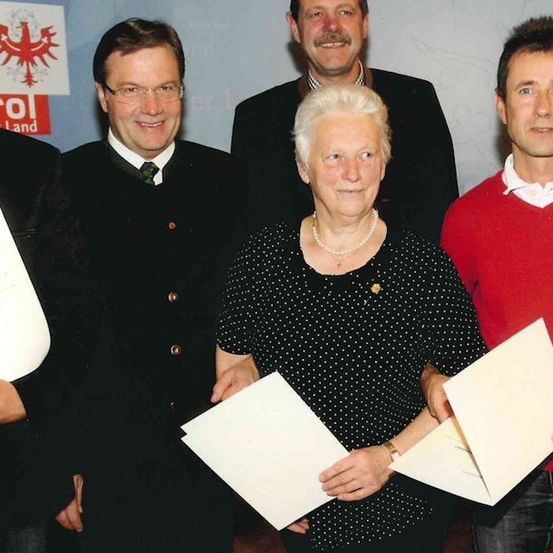 Fünf Personen stehen lächelnd zusammen und halten Urkunden. Eine Frau in der Mitte trägt eine gepunktete Bluse und eine Halskette. Dahinter stehen drei Männer in Anzügen und einer in einem roten Hemd.