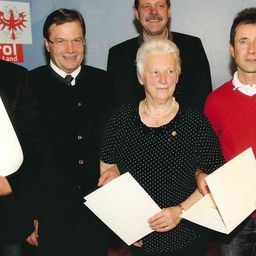 Fünf Personen stehen lächelnd zusammen und halten Urkunden. Eine Frau in der Mitte trägt eine gepunktete Bluse und eine Halskette. Dahinter stehen drei Männer in Anzügen und einer in einem roten Hemd.