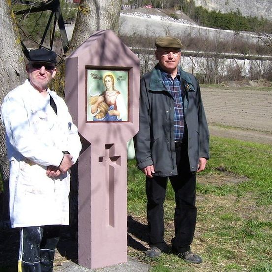 Zwei Männer stehen neben einem bemalten Holzkreuz auf einem Feld mit Bäumen und einer Mauer im Hintergrund.