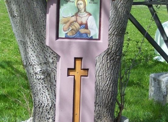 Eine rosafarbene Holztafel mit einem religiösen Bild und einem Kreuz ist an einem Baumstamm in einem Grasbereich befestigt.