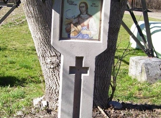 Ein Betonkreuz mit einem Bild einer Frau, die ein Baby hält, ist an einem Baumstamm befestigt. Das Kreuz steht auf einem Rasen.