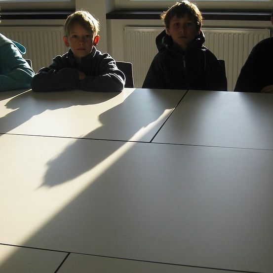 Vier Jungen sitzen um einen Tisch herum, ihre Schatten fallen auf die Oberfläche. Sie scheinen sich in einem Klassenzimmer zu befinden.