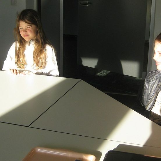 Zwei junge Mädchen sitzen an einem Tisch, eine schaut in die Kamera, während die andere wegschaut, mit Sonnenlicht, das die Szene beleuchtet.
