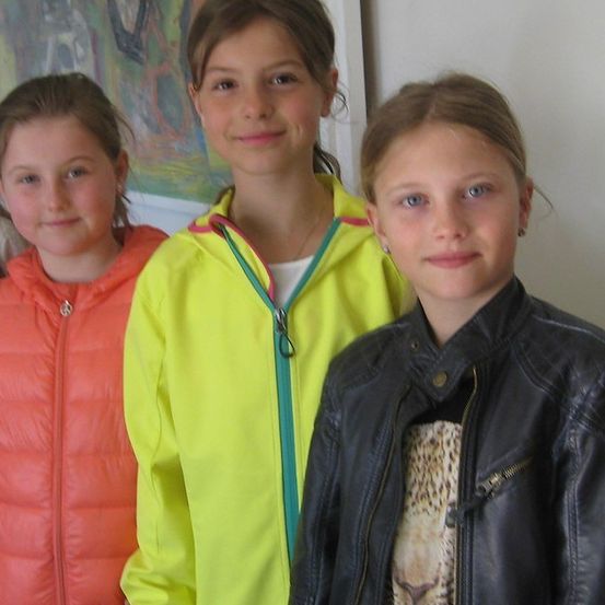 Vier Mädchen stehen zusammen und lächeln für ein Foto. Das Mädchen auf der linken Seite trägt eine rote Jacke, das zweite Mädchen eine gelbe Jacke und das dritte Mädchen eine schwarze Lederjacke. Das vierte Mädchen ist teilweise sichtbar.