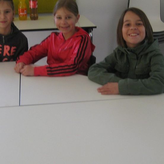 Drei Mädchen sitzen an einem Tisch und lächeln für ein Foto. Das Mädchen auf der linken Seite trägt eine schwarze Kapuzenjacke mit dem Wort 'HIER'. Das Mädchen in der Mitte trägt eine rote Jacke und das Mädchen auf der rechten Seite eine grüne Jacke. Hinter ihnen steht ein Tisch mit zwei Flaschen.