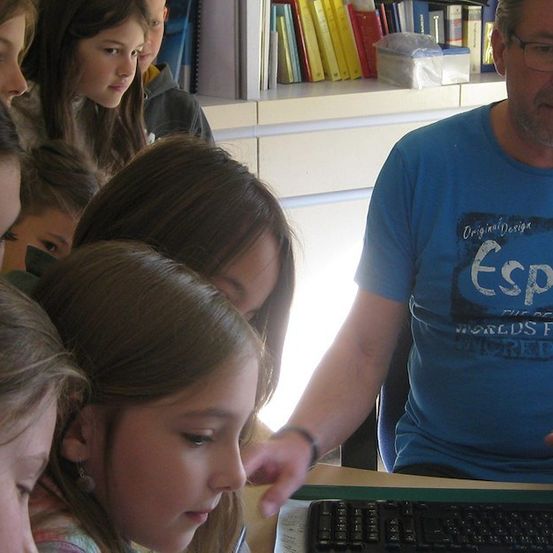 Ein Mann in einem blauen T-Shirt mit dem Wort 'Esprit' unterrichtet eine Gruppe junger Mädchen in einem Klassenzimmer. Sie schauen auf einen Computerbildschirm.