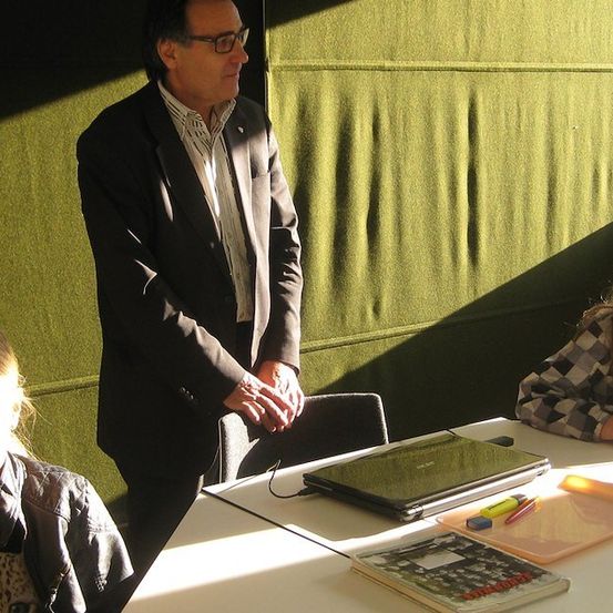 Ein Mann im Anzug steht hinter einem Schreibtisch mit zwei Mädchen. Ein Mädchen lächelt und das andere schaut den Mann an. Der Schreibtisch hat einen Laptop, Bücher und andere Gegenstände.