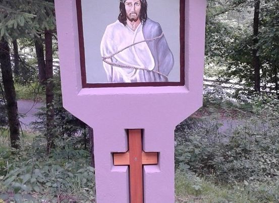 Ein lilafarbener Pfosten mit einem hölzernen Kreuz unten und einem Bild von Jesus Christus in Ketten darauf.
