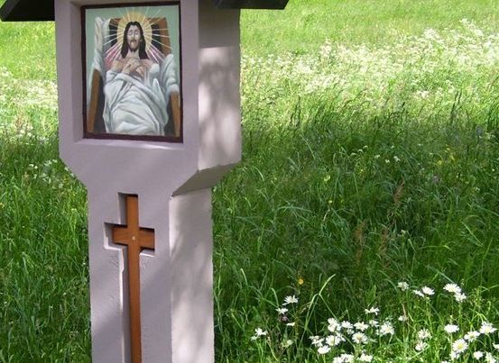 Ein kleiner Betonkreuz mit einem Bild von Jesus darauf, in einem Grasfeld mit weißen Blumen.