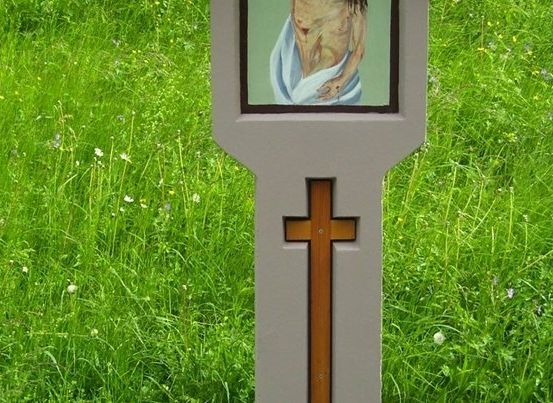 Ein hölzernes Kreuz mit einer kleinen Malerei eines Mannes, der in ein weißes Tuch gewickelt ist, steht auf einer Wiese.
