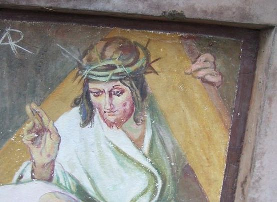 Ein verblasstes Gemälde von Jesus Christus am Kreuz, mit Dornenkrone, Friedenszeichen in der Hand und einem Aufkleber 'AR' oben links.