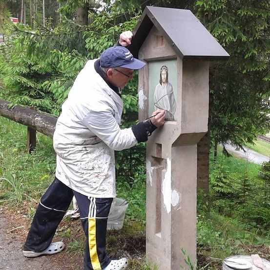 Ein Mann mit blauer Mütze und Brille malt ein religiöses Bild auf eine Betonstruktur am Straßenrand.