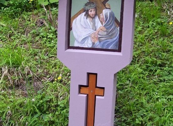 Ein lila Kreuz mit einem religiösen Bild von Jesus und einer Frau darauf. Das Kreuz steht auf einem Grasfeld.